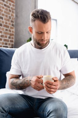 Beyaz tişörtlü ve kot pantolonlu dalgın genç bir adam bir fincan çayla yatakta oturuyor.