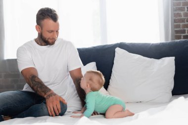 young tattooed man in white t-shirt and jeans sitting near infant son crawling on bed in baby romper clipart