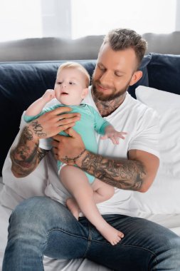 Kotlu genç sakallı adam yatakta bebek kucağında oturuyor.