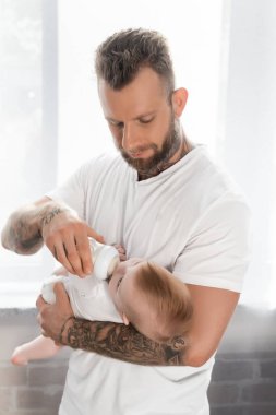 Genç dövmeli bir adam pencerenin yanında dururken bebeğini biberonla besliyor.