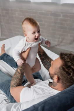 Yatakta otururken heyecanlı bir çocuğu tutan dövmeli genç bir babanın seçici odağı.