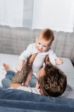 overhead view of tattooed man holding excited infant son while sitting in bed clipart