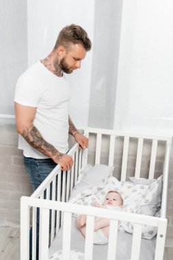 Genç dövmeli adam beşiğin yanında dururken bebeğe bakıyor.