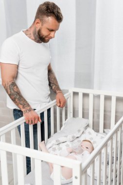 young tattooed man looking at infant kid lying in crib with baby bottle clipart