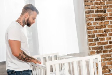 Evde beşiğin yanında duran dövmeli genç adamın yan görüntüsü