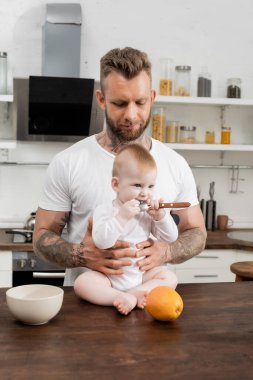 Küçük çocuk annesinin yanında kaşık tutarken mutfak masasında genç babasının yanında oturuyor.
