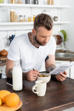 Mutfakta kahvaltı ederken gazete okuyan sakallı, dövmeli bir adamın seçici odak noktası.