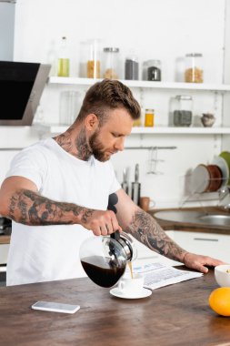 Dövmeli beyaz tişörtlü adam gazete okuyor ve mutfak masasında cep telefonunun yanına kahve döküyor.