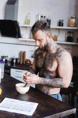 Genç dövmeli bir adam kahvaltıda kasenin yanında dururken akıllı telefondan sohbet ediyor.