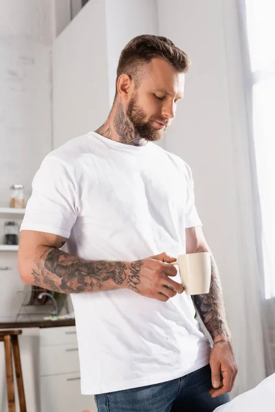 お茶を飲みながら台所に立つ白いTシャツを着た思慮深い若者 — ストック写真