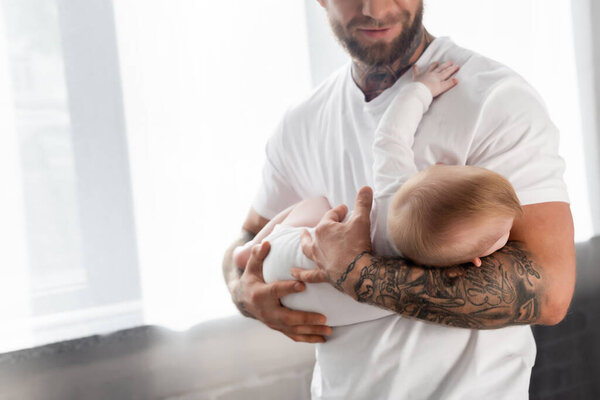 selective focus of young tattooed man in white t-shirt holding infant son at home