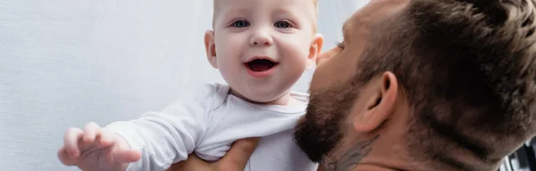 興奮した赤ちゃんを抱える父親の選択的焦点口を開けてカメラを見るパノラマの概念 — ストック写真