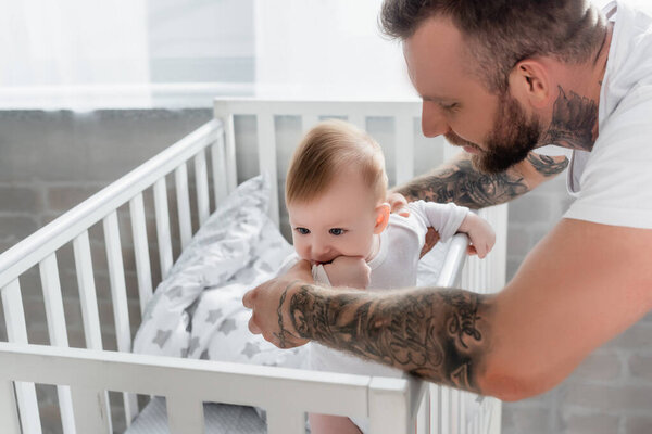 young father supporting infant son standing in crib with hand in mouth
