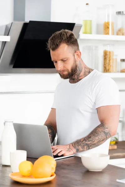 Young Tattooed Freelancer Using Laptop Bowl Oranges Bottle Milk Kitchen — Stock Photo, Image