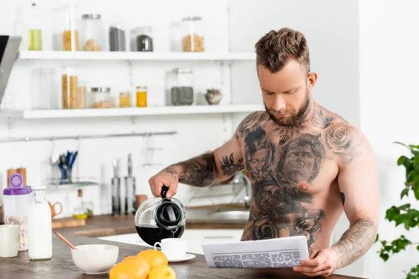 young tattooed man pouring coffee and reading newspaper during breakfast in kitchen