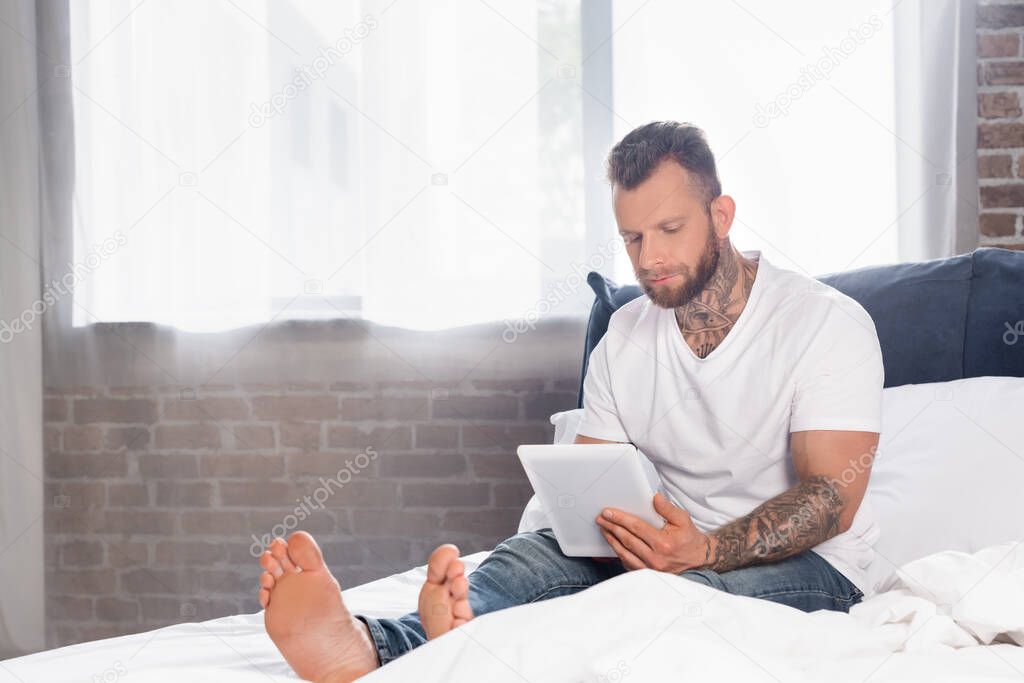 tattooed man in white t-shirt sitting in bed near window and using digital tablet