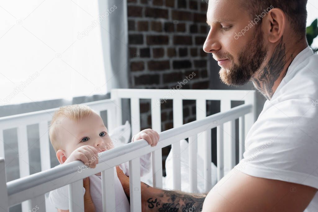 bearded, tattooed man supporting infant kid standing in cot