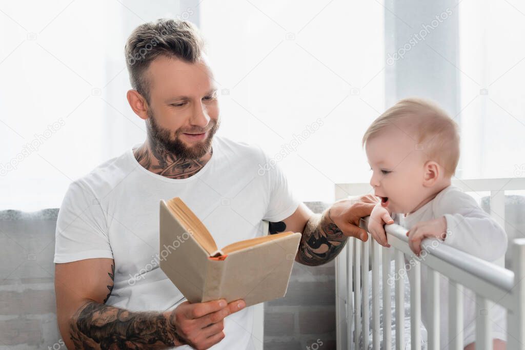young tattooed man reading book to excited infant boy standing in crib