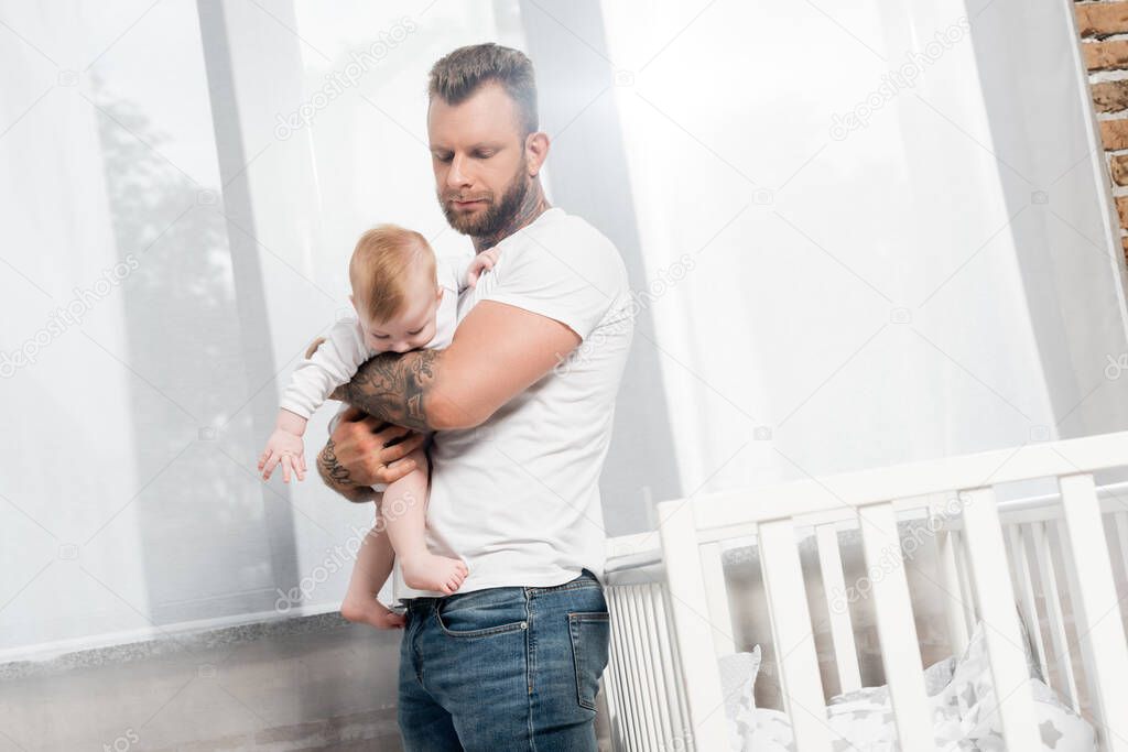 tattooed man holding baby boy while standing near crib at window