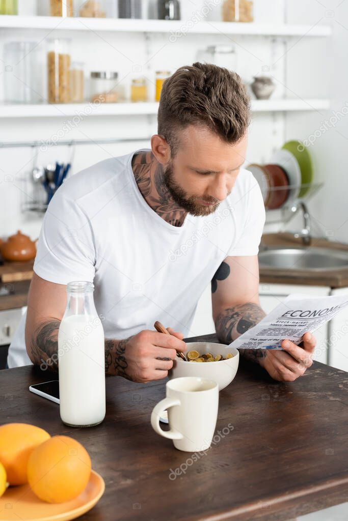 selective focus of bearded, tattooed man reading newspaper while having breakfast in kitchen