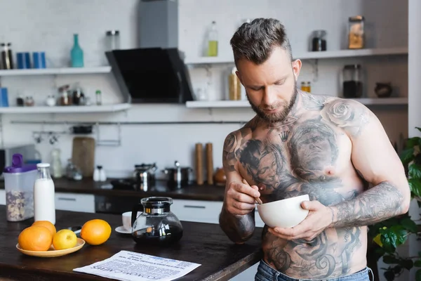 muscular, tattooed man holding bowl with breakfast while standing in kitchen