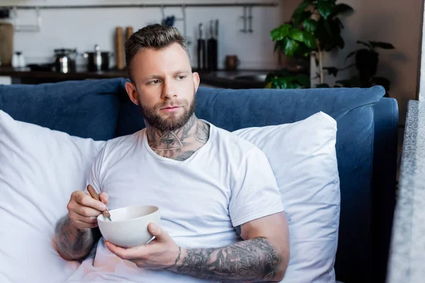 Νεαρός Άνδρας Τατουάζ Κοιτάζει Αλλού Ενώ Παίρνει Πρωινό Στην Κρεβατοκάμαρα — Φωτογραφία Αρχείου