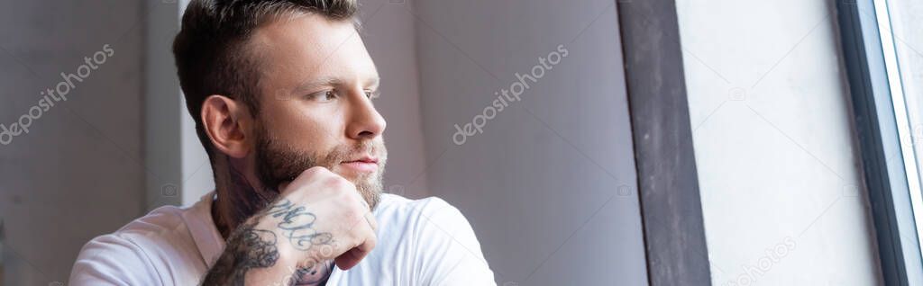 horizontal concept of pensive tattooed man holding hand near face while looking away at home