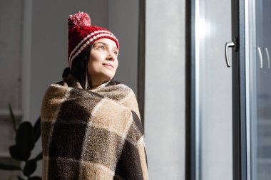 dreamy woman in knitted hat, wrapped in warm plaid blanket, looking away while standing near window at home clipart