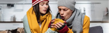panoramic crop of boyfriend in knitted scarf and hat warming hands of girlfriend at home clipart