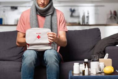 partial view of ill man with first aid kit sitting on sofa near medicines clipart