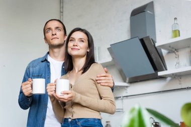 Genç çift mutfakta durup gözlerini kaçırırken ellerinde kahve fincanları tutuyorlar.
