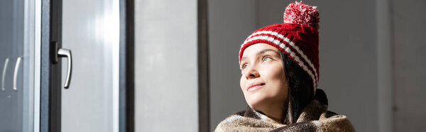 website header of young woman in warm knitted hat looking away while standing near window at home