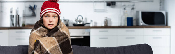 horizontal concept of cold woman in warm hat, wrapping in warm plaid blanket while sitting in kitchen