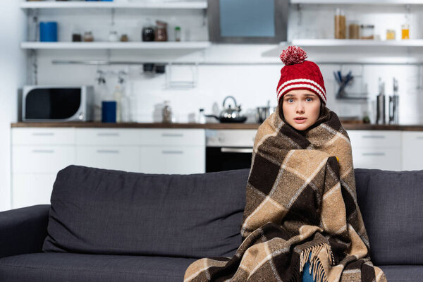 freezing woman in knitted hat, covering with warm plaid blanket while sitting on couch in cold kitchen