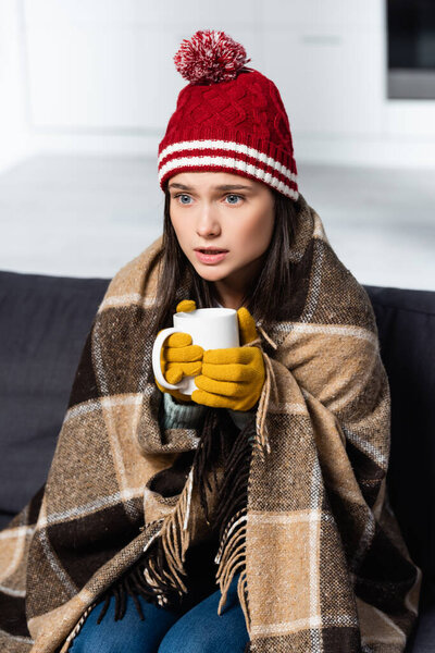 cold woman, wrapped in plaid blanket, wearing knitted hat and gloves, holding warming drink in kitchen