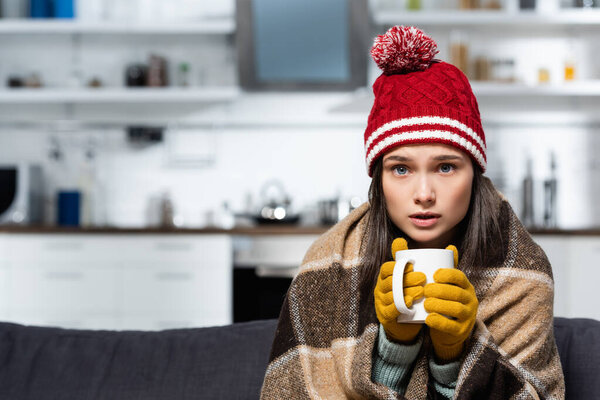 freezing woman, wrapped in plaid blanket, wearing knitted gloves and hat, holding warm tea in cold kitchen