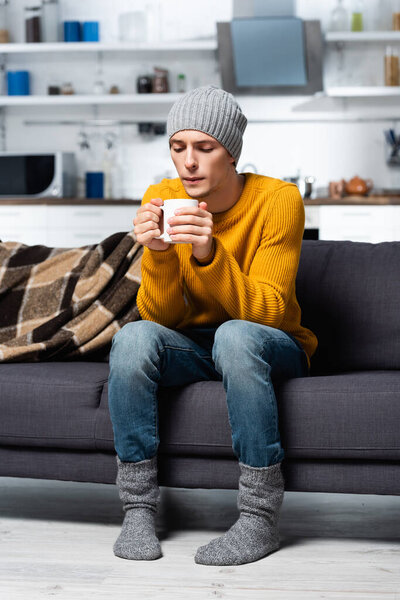 young man in knitted sweater, hat and socks blowing on hot tea while sitting on sofa in cold kitchen