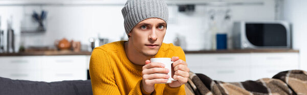 panoramic orientation of young man in knitted sweater and hat looking at camera while holding cup of warming beverage