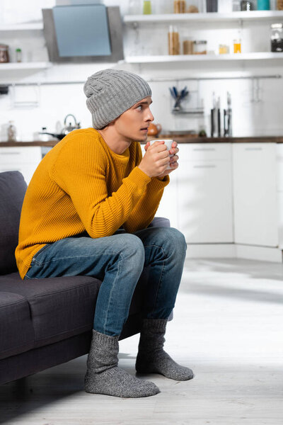 freezing man in knitted sweater, hat and socks holding cup of warm beverage in kitchen