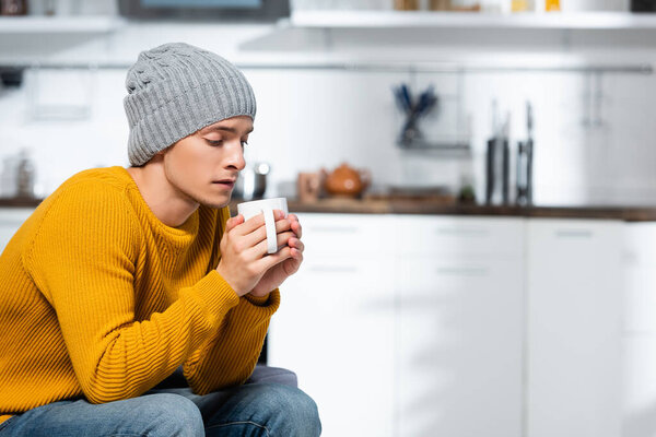 young man in knitted sweater and hat holding cup of warm tea in kitchen