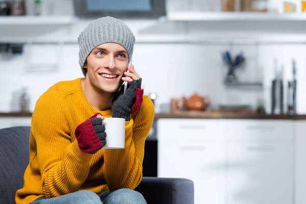 joyful man in fingerless gloves and knitted hat talking on cellphone while holding cup of warm drink 