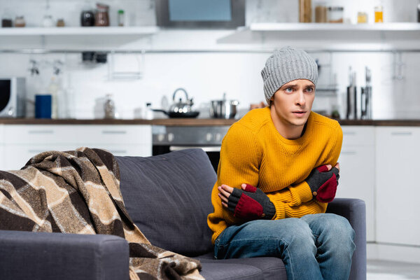 Молодой человек в теплой шляпе и перчатках без пальцев обнимает себя, сидя на диване на холодной кухне