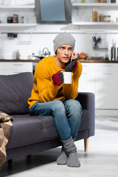freezing man in warm hat, socks and fingerless gloves talking on mobile phone while holding cup of tea in kitchen