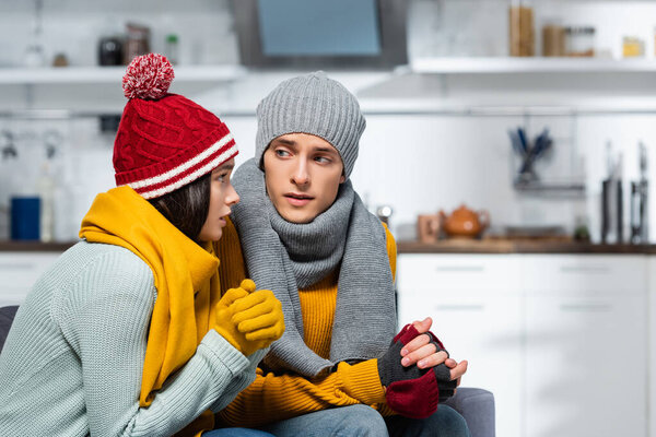 young couple in knitted hats, scarfs and gloves looking at each other while freezing in cold kitchen