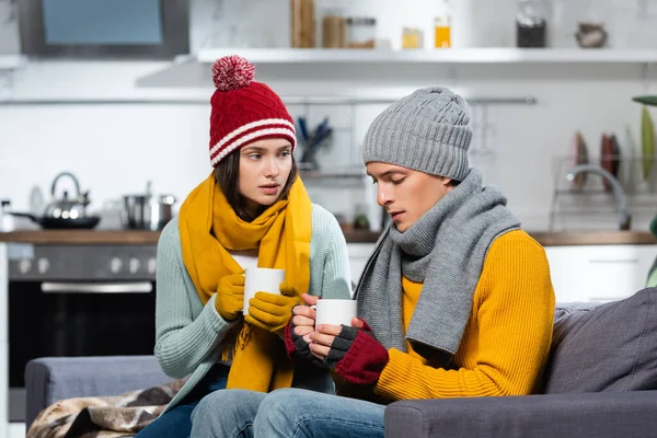 在寒冷的厨房里 戴着针织帽子 戴着围巾 戴着手套 手里拿着热饮的冰冻夫妇 — 图库照片