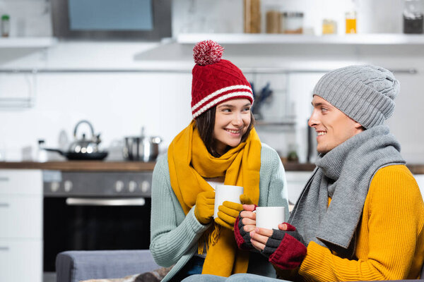 joyful couple in warm hats, scarfs and gloves looking at each other while holding cups of warming beverage in kitchen