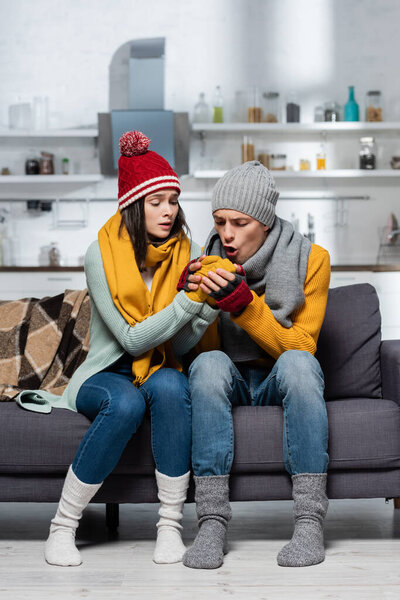 young man in knitted hat and scarf warming hands of girlfriend while sitting on sofa in cold kitchen