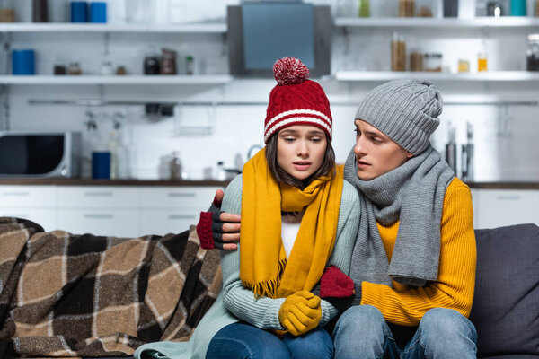 young man in warm hat and scarf hugging freezing girlfriend while sitting in cold kitchen