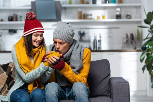 young man in knitted hat, scarf and gloves warming hands of freezing girlfriend