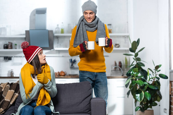 cold woman in knitted hat and gloves looking at boyfriend carrying warm tea in kitchen
